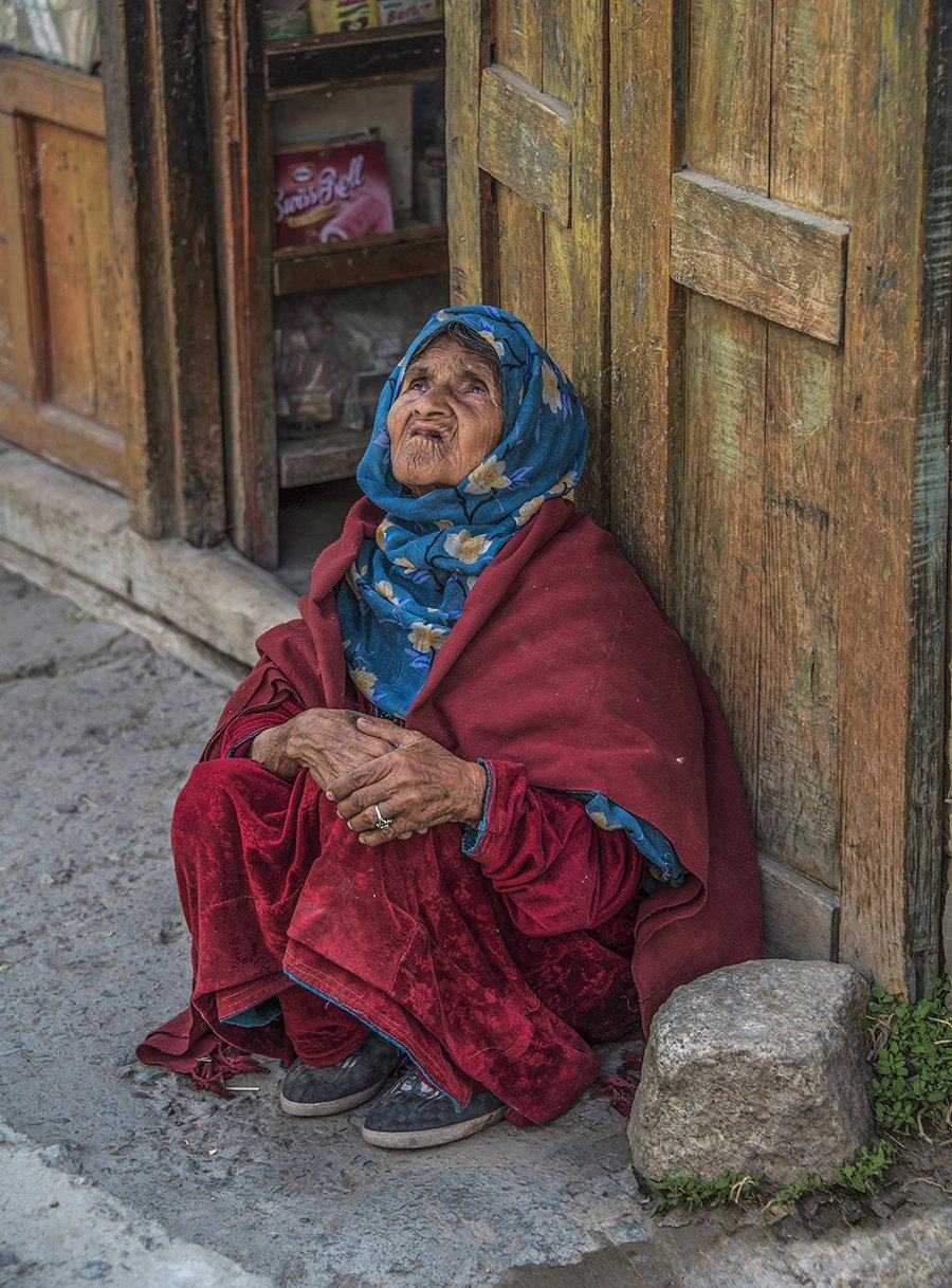 Elderly woman in a red shawl and blue headscarf sitting by a wooden door, looking up