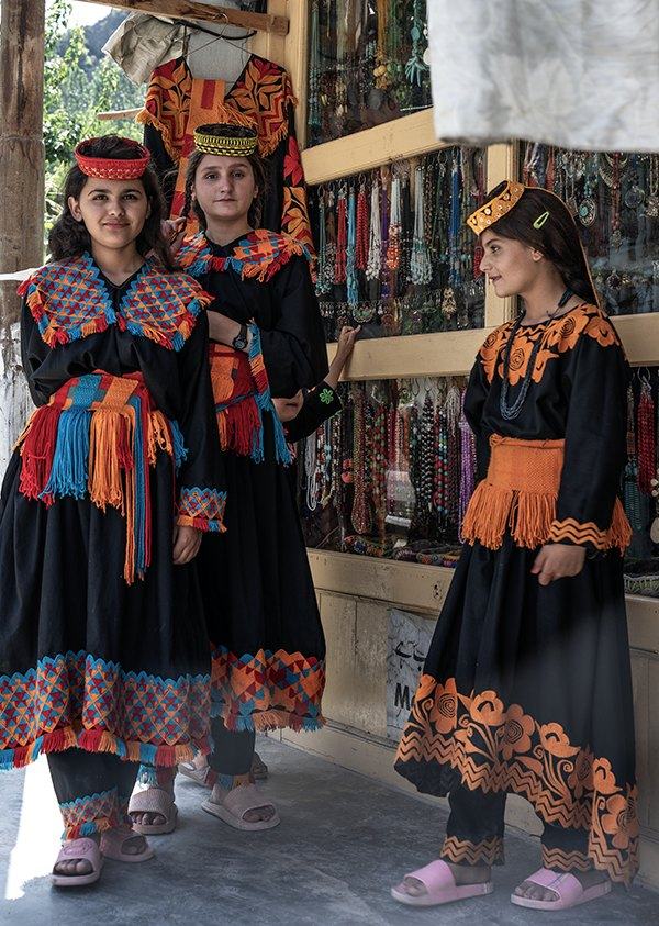Three girls in black dresses with orange embroidery standing outside a bead shop