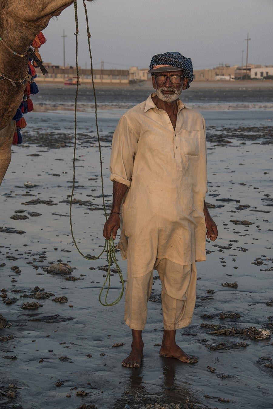Man in pale clothes standing barefoot in shallow water at dusk beside a camel