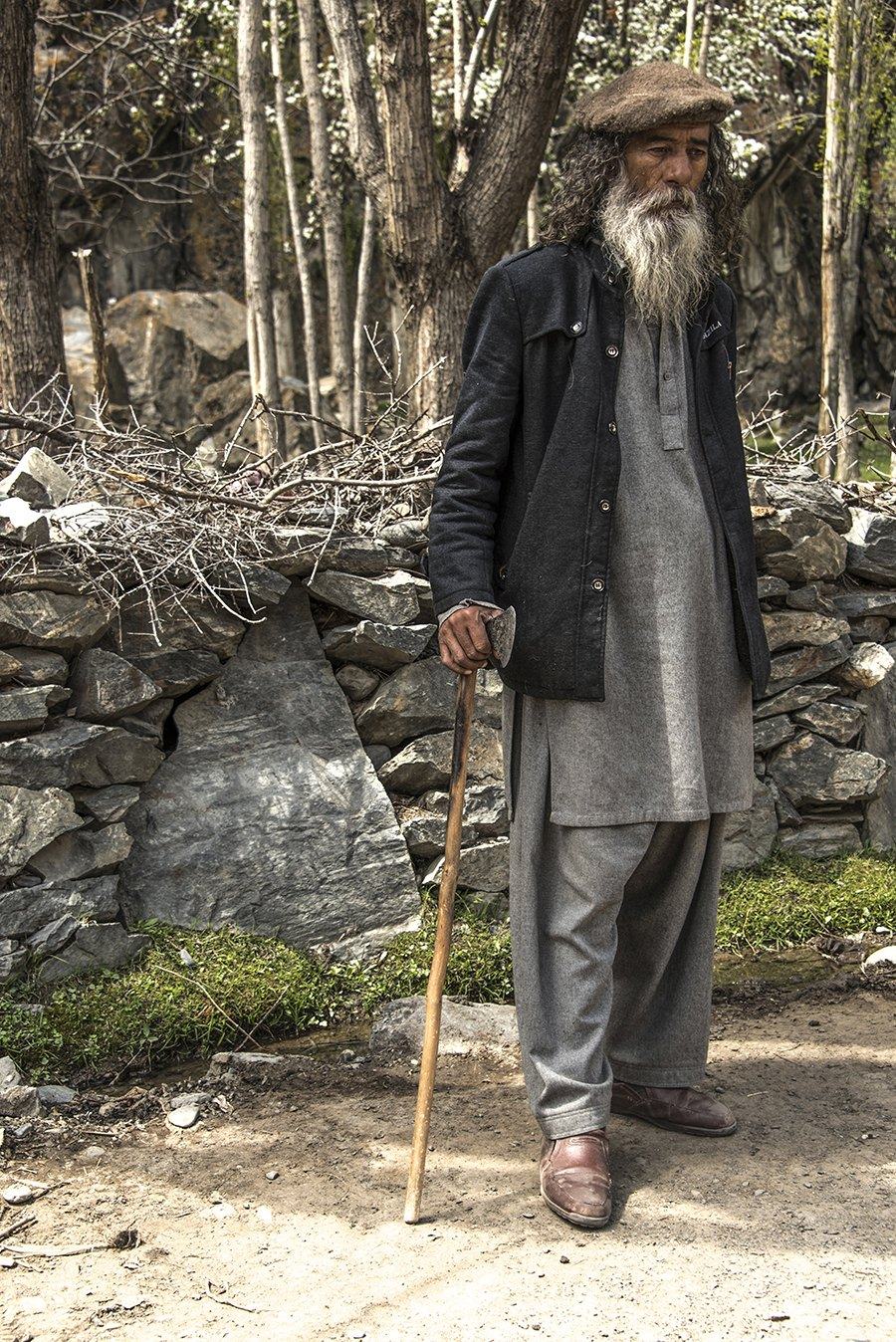 Bearded man in a flat cap and long coat leaning on a walking stick by a stone wall